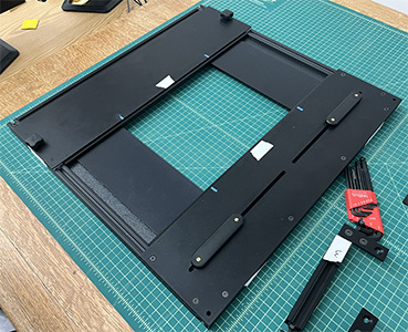 A square table made of black aluminum and wood. The four legs have been removed, and there is an empty space in the center of the table. There are two rubberized knobs on the end farthest the camera, and two low grips on the side near the camera. Nest to the table is one of the removed legs and a set of hex keys.