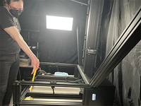 An image of the 413 copy stand from the side. The tabletop has been removed, showing the empty space below the top, one of the lights, and the center column. A woman in black wearing a dust mask is cleaning the copy stand with a swiffer duster.