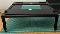 A square table made of black aluminum and wood. It has four legs, and there is an empty space in the center of the table. There are two rubberized knobs on the end nearest the camera, and two low grips on the side away from the camera.