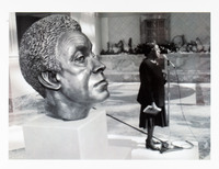 Black and white photo of a bust of Brooks located foreground left and Brooks located background right speaking into a microphone.