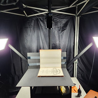 A front-facing view of the 413 copy stand. It is a flat table with two aluminum stands coming off the back at 45 degrees and a central vertical column in the center of the back. The aluminum stands hold two LED lights facing in to the table. There is a medium-format camera mounted on the column pointing down at the tabletop. There is a book strapped to the cradle underneath the camera.